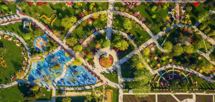 Geometrik yürüyüş yolları, renkli çiçek tarhları ve mavi zeminli çocuk oyun alanı içeren büyük şehir parkının kuşbakışı görünümü