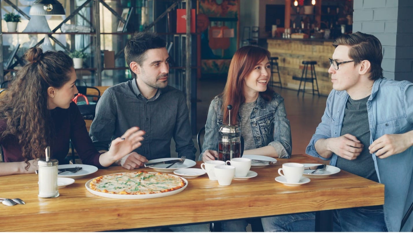 Kafede ahşap bir masada pizza yiyip kahve içerek sohbet eden dört arkadaş